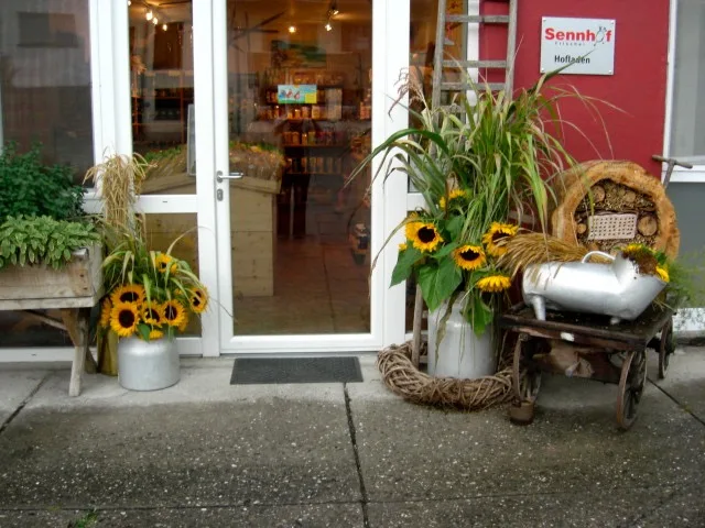 Eingang des Sennhof Hofladens mit rotem Schild, Sonnenblumen in Milchkannen und einem Leiterwagen