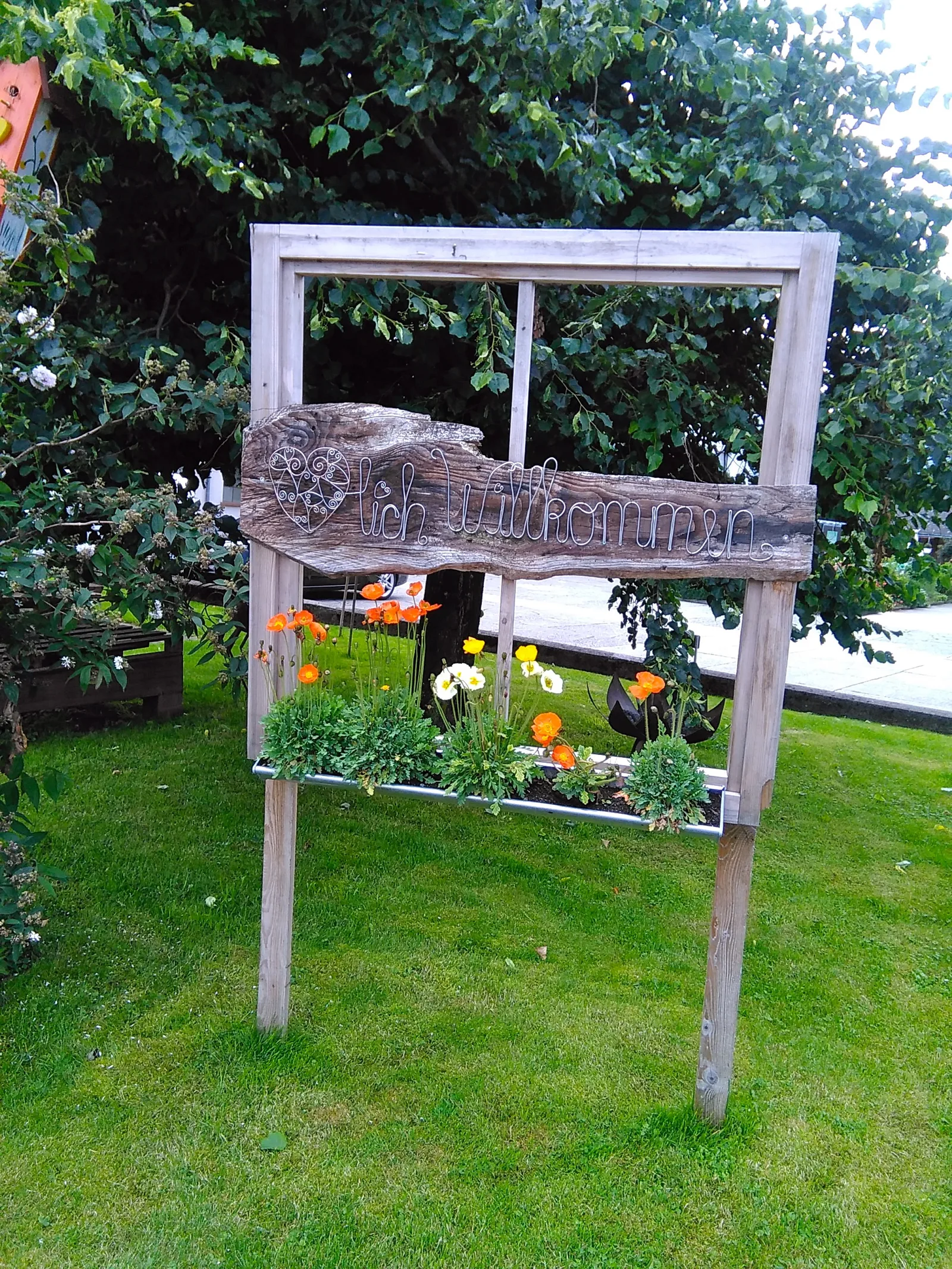 Alter Fensterrahmen im Gras beim Hof, darauf in Draht geschrieben Herzlich Willkommen, davor Mohnblumen